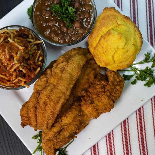 Fried catfish fillets, baked beans, spaghetti, and cornbread on a white plate with a striped napkin.