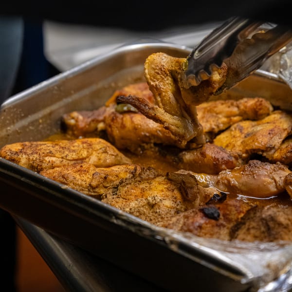 Tongs lift a piece of seasoned, cooked chicken from a large pan of Detroit Soul food.