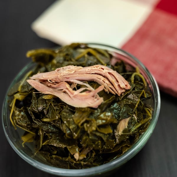 A glass bowl of Detroit Soul's collard greens, topped with shredded meat, on a dark surface.