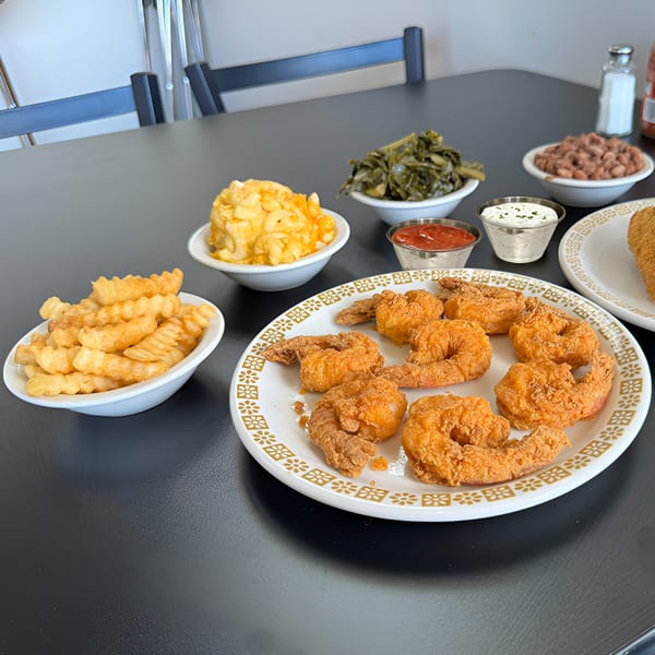 Detroit Soul's fried shrimp, crinkle-cut fries, mac and cheese, collard greens, and beans on a black table.